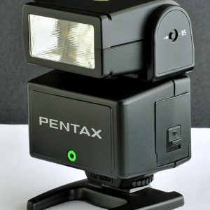 Könnte beinhalten: Ein schwarzer Pentax-Blitz mit eingebautem Blitzlicht und schwenkbarem Kopf. Der Blitz verfügt über eine grüne LED-Anzeige und einen kleinen schwarzen Knopf an der Seite.