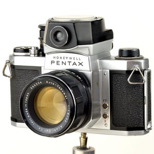 Może przedstawiać: Zabytkowy aparat fotograficzny Honeywell Pentax w srebrno-czarnej obudowie. Obiektyw z napisem "Auto-Takumar 1:1.8/55". Widoczna nazwa marki "Honeywell Pentax". Aparat zamontowany na statywie.