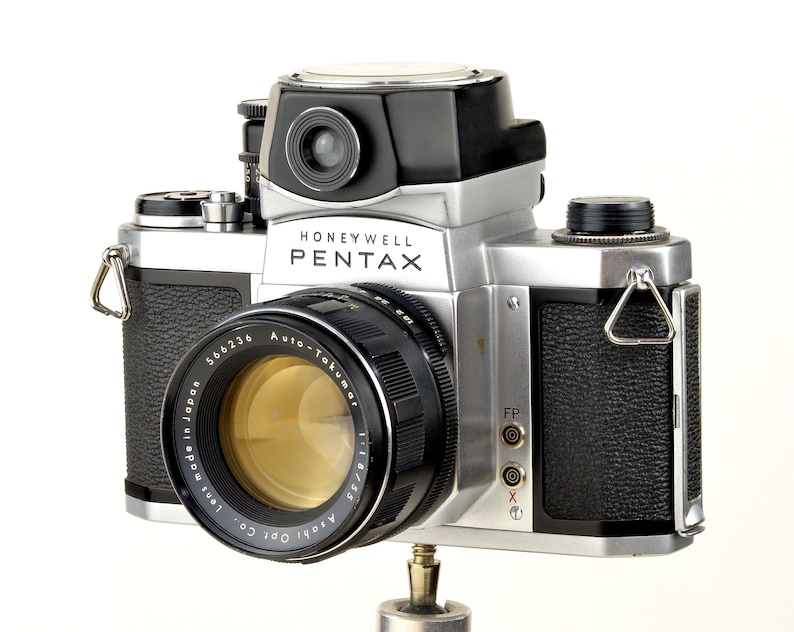 Może przedstawiać: Zabytkowy aparat fotograficzny Honeywell Pentax w srebrno-czarnej obudowie. Obiektyw z napisem "Auto-Takumar 1:1.8/55". Widoczna nazwa marki "Honeywell Pentax". Aparat zamontowany na statywie.