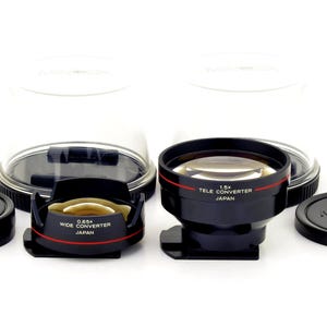 May include: Two black camera lens converters, one labeled 0.85x Wide Converter Japan and the other 1.5x Tele Converter Japan, each with a red accent line. Includes two black Minolta lens caps and two clear plastic protective dome covers.