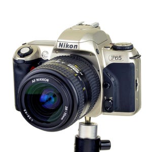May include: A silver and black Nikon F65 camera with a 28-70mm lens attached. The camera is mounted on a black tripod. The lens has a green and blue reflection.
