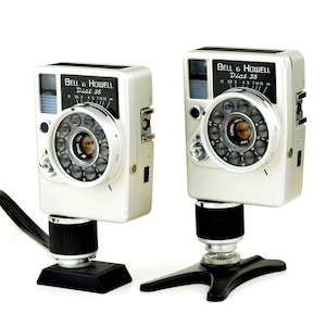 May include: Two silver Bell & Howell Dial 35 cameras with white bodies and black accents. Each camera features a circular lens assembly with a black dial, a black rectangular plate with white text, and is mounted on a black camera stand base.