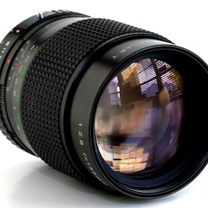 May include: A black camera lens with a silver ring and a black rubber grip. The lens has a depth of field scale and a focus ring. The lens is marked "HART 1:2.8 f=135mm".