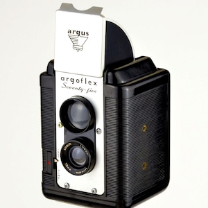 May include: Vintage Argoflex Seventy-five camera. The black and silver camera has two lenses and a viewfinder. The camera has the Argus logo and the text "Argoflex Seventy-five". The camera is mounted on a tripod.
