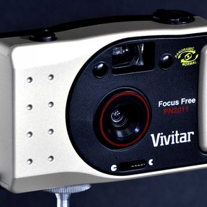 May include: A silver and black Vivitar Focus Free PN2011 camera with a red lens ring. The camera has a panoramic setting and a focus free lens.