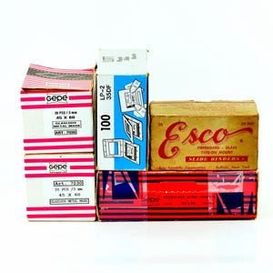 Può includere: Una collezione di cinque scatole rettangolari per telaietti per diapositive. Due scatole bianche con strisce rosa riportano la scritta Gepe 20 pcs 3 mm 45 x 60. Una scatola blu indica LP-2 35DF 100. Una scatola marrone riporta Esco 20 35 mm Slide Binders.