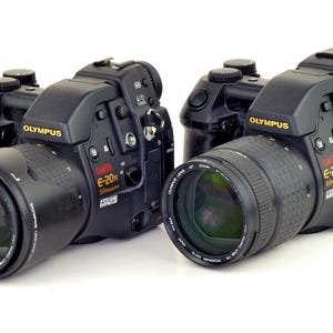 May include: Two black Olympus E-20N digital cameras positioned side by side. Each camera features a prominent lens, a textured grip, and various control buttons on the top and side. The brand name Olympus and the model E-20N are printed in yellow and red.