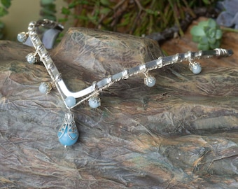 May include: A silver headband with a delicate design and blue gemstone accents. The headband has a V-shape and a dangling gemstone pendant.