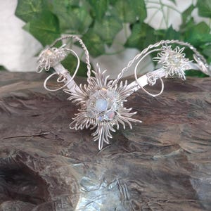 May include: A silver-colored, handcrafted headpiece with a central floral design and delicate wirework. The headpiece features a symmetrical design with spiraling accents and small, sparkling embellishments. The piece is displayed on a textured, dark brown surface.