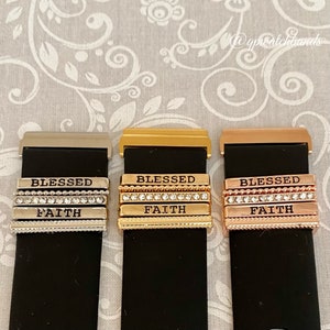K&ouml;nnte beinhalten: Drei Uhrenarmb&auml;nder mit austauschbaren Charms. Die Charms sind silber, gold und ros&eacute;gold und haben die Worte "Blessed" und "Faith" eingraviert. Die Charms sind mit kleinen Strasssteinen verziert.