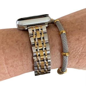 Può includere: Cinturino per orologio in metallo argentato e dorato con bracciale a rete. Il cinturino è attaccato a un quadrante argentato. Il cinturino ha una combinazione di maglie argentate e dorate. Il bracciale a rete ha dettagli dorati.