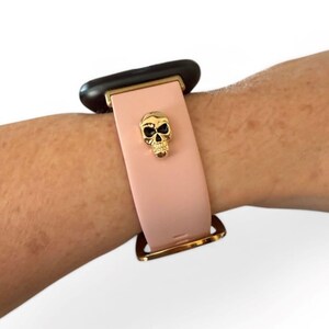 Op de afbeelding: Een lichtroze smartwatch bandje met een goudkleurige schedelbedel. De band heeft een goudkleurige gesp en een zwarte wijzerplaat. De band wordt getoond op een pols.