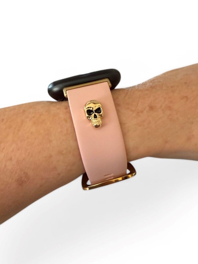 Op de afbeelding: Een lichtroze smartwatch bandje met een goudkleurige schedelbedel. De band heeft een goudkleurige gesp en een zwarte wijzerplaat. De band wordt getoond op een pols.