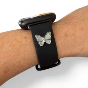 May include: A black smartwatch band with a silver butterfly charm. The butterfly has a white center and is outlined with small, clear crystals. The watch face is dark with a gold-colored trim.