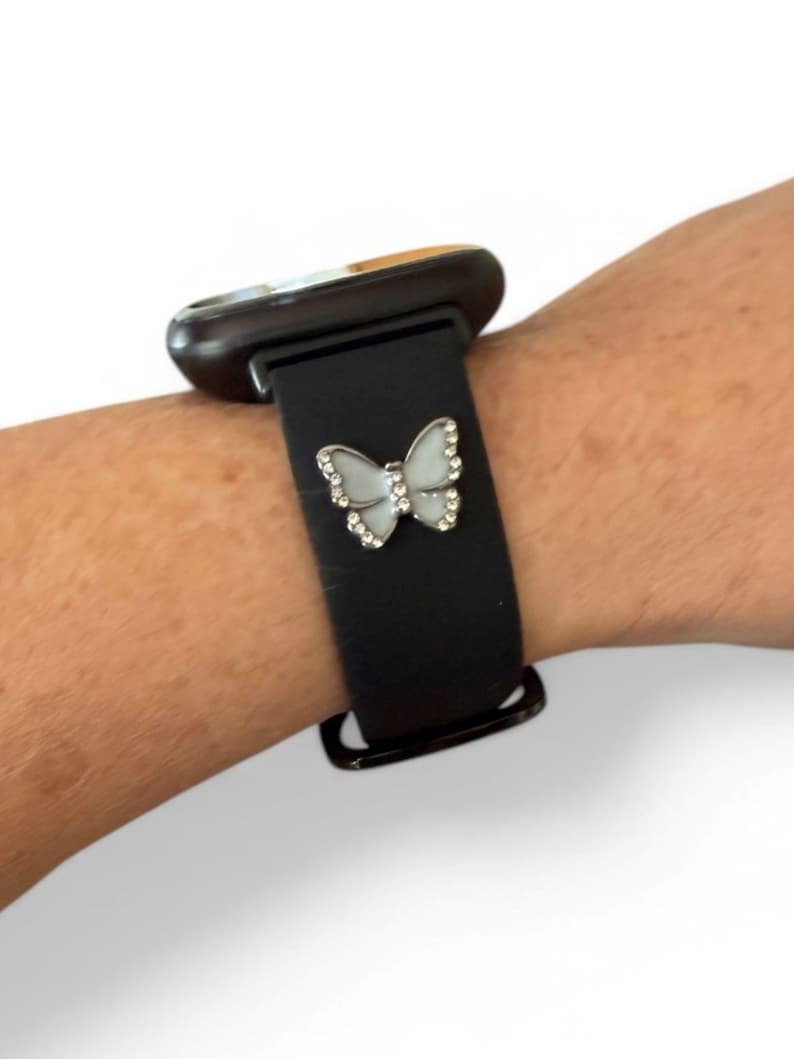 May include: A black smartwatch band with a silver butterfly charm. The butterfly has a white center and is outlined with small, clear crystals. The watch face is dark with a gold-colored trim.