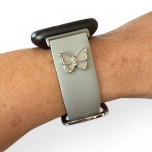May include: A gray smartwatch band with a silver butterfly charm. The butterfly is encrusted with small, clear crystals. The band is attached to a black smartwatch face and has a silver buckle.