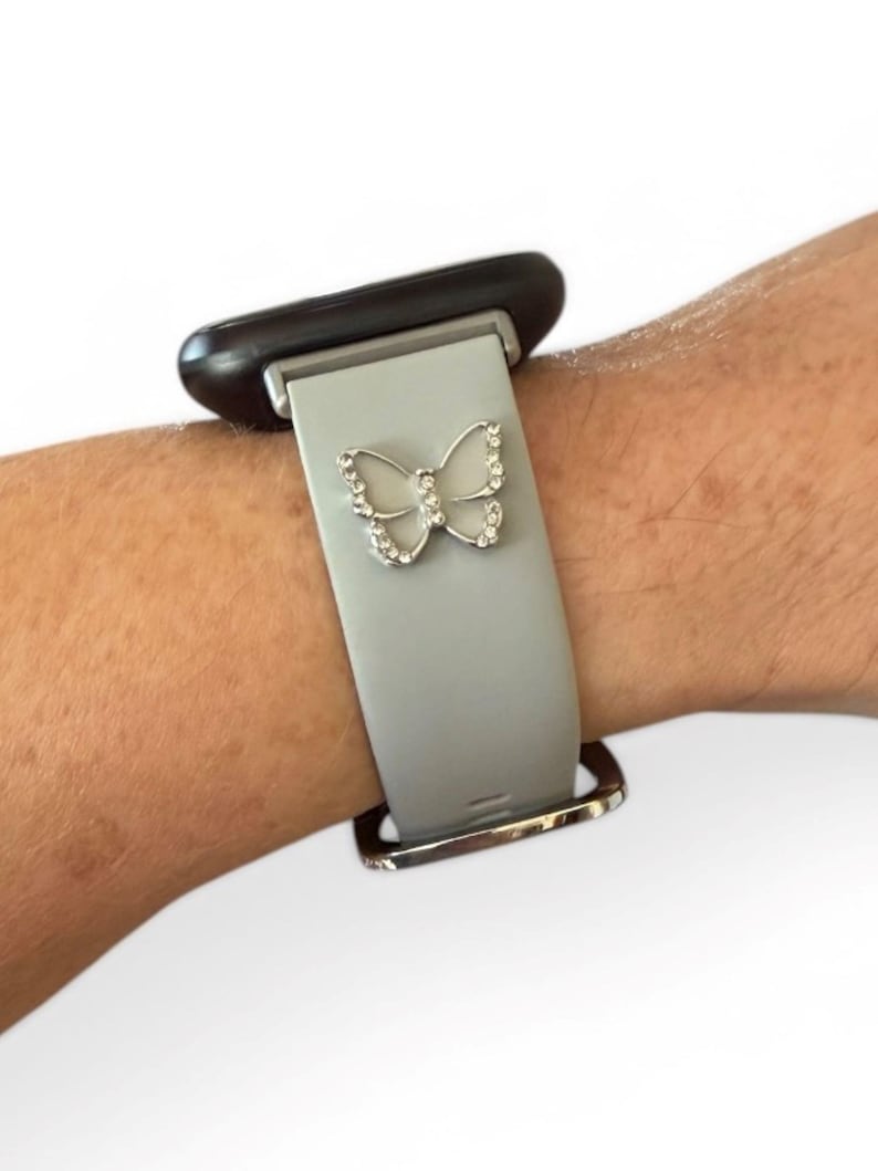 May include: A gray smartwatch band with a silver butterfly charm. The butterfly is encrusted with small, clear crystals. The band is attached to a black smartwatch face and has a silver buckle.