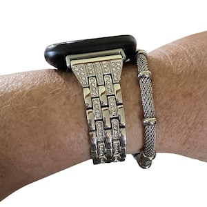 May include: A silver-toned watch band with a rectangular design, embellished with clear rhinestones. The watch band is connected to a black smartwatch. A silver mesh bracelet is next to the watch band.