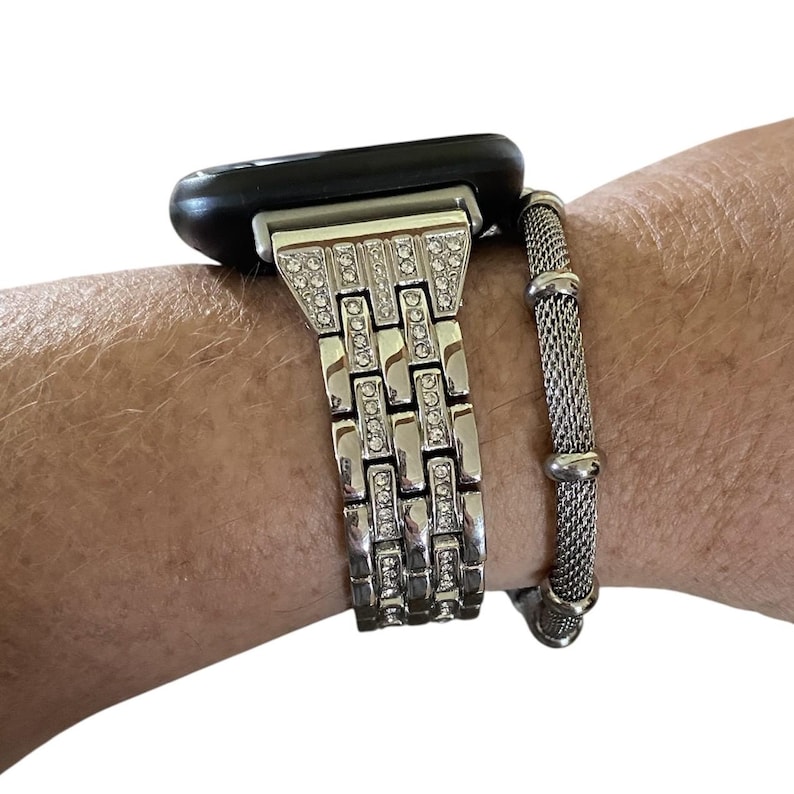 May include: A silver-toned watch band with a rectangular design, embellished with clear rhinestones. The watch band is connected to a black smartwatch. A silver mesh bracelet is next to the watch band.
