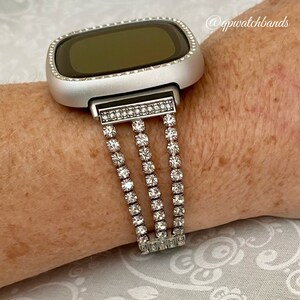 K&ouml;nnte beinhalten: Silberne Smartwatch mit einem durchsichtigen Kristallband. Das Band hat drei Reihen mit Kristallen.