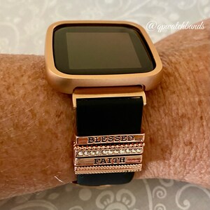 K&ouml;nnte beinhalten: Eine ros&eacute;goldene Smartwatch mit einem schwarzen Silikonarmband und austauschbaren Charms. Die Charms sind mit den Worten "Blessed" und "Faith" graviert.