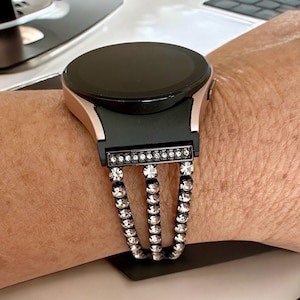 Pode incluir: Um smartwatch preto com uma banda de ouro rosa e uma pulseira incrustada de cristais. A pulseira é feita de três fileiras de cristais.