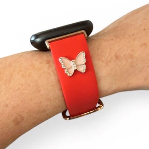 May include: Red smartwatch band with a gold-colored clasp and a decorative butterfly. The butterfly is embellished with small, clear crystals and a pink center. The watch face is black.
