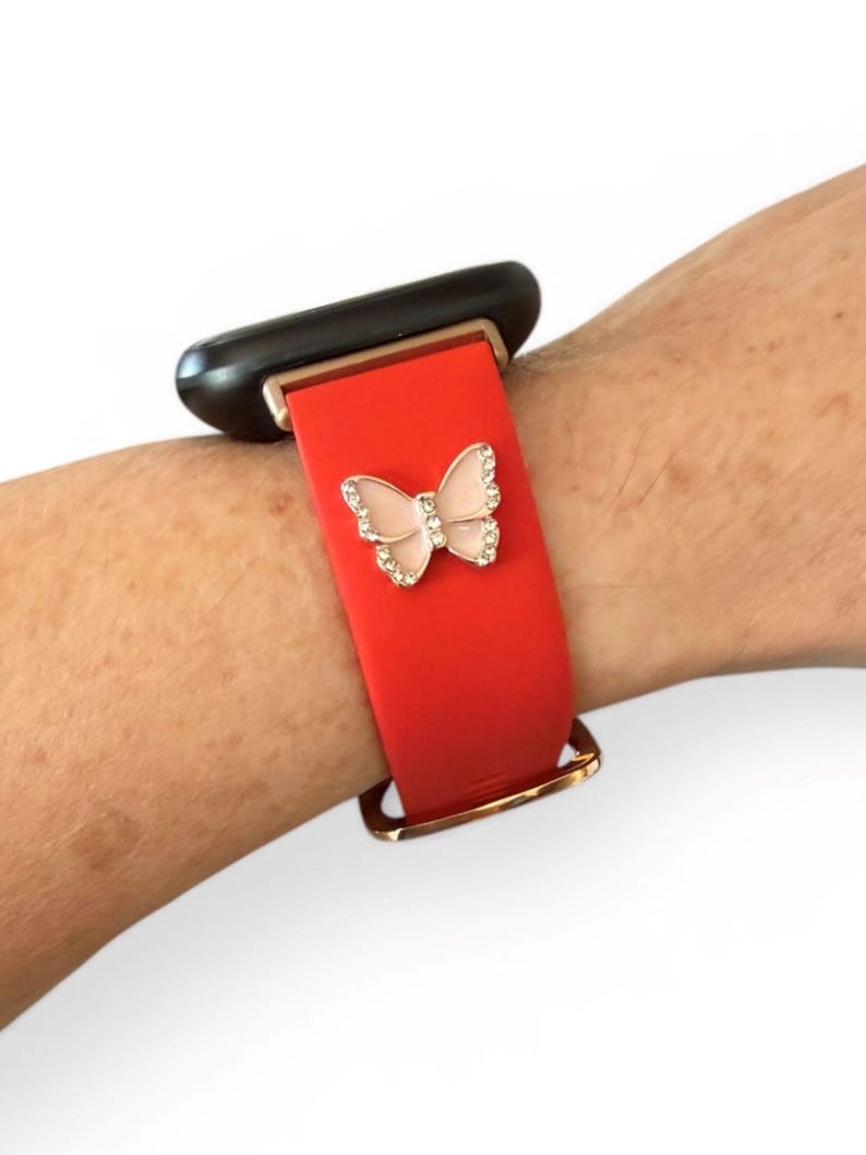 May include: Red smartwatch band with a gold-colored clasp and a decorative butterfly. The butterfly is embellished with small, clear crystals and a pink center. The watch face is black.