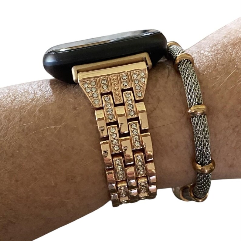 May include: Rose gold watch band with sparkling crystal accents. The band is connected to a black smartwatch. A silver and gold bracelet is also visible.