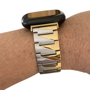 K&ouml;nnte beinhalten: Ein zweifarbiges Uhrenarmband mit schwarzem Zifferblatt. Das Armband wechselt zwischen gold- und silberfarbenen Metallgliedern. Das Zifferblatt hat eine dunkelbraune Farbe.