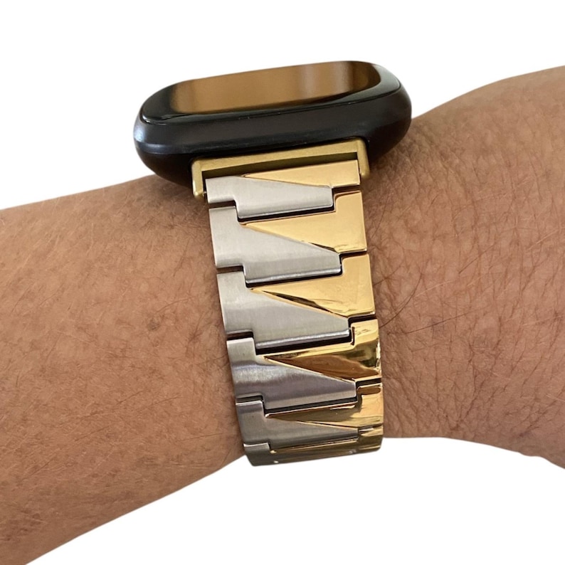 K&ouml;nnte beinhalten: Ein zweifarbiges Uhrenarmband mit schwarzem Zifferblatt. Das Armband wechselt zwischen gold- und silberfarbenen Metallgliedern. Das Zifferblatt hat eine dunkelbraune Farbe.