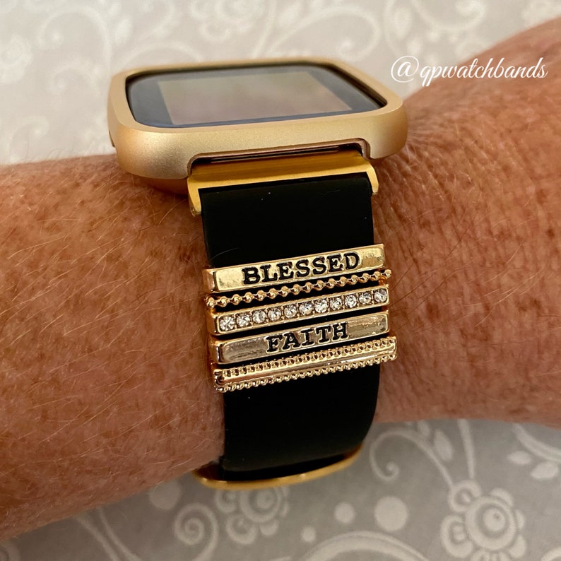 K&ouml;nnte beinhalten: Goldene Smartwatch mit schwarzem Silikonarmband und goldenen Charms mit den Aufschriften "Blessed" und "Faith".