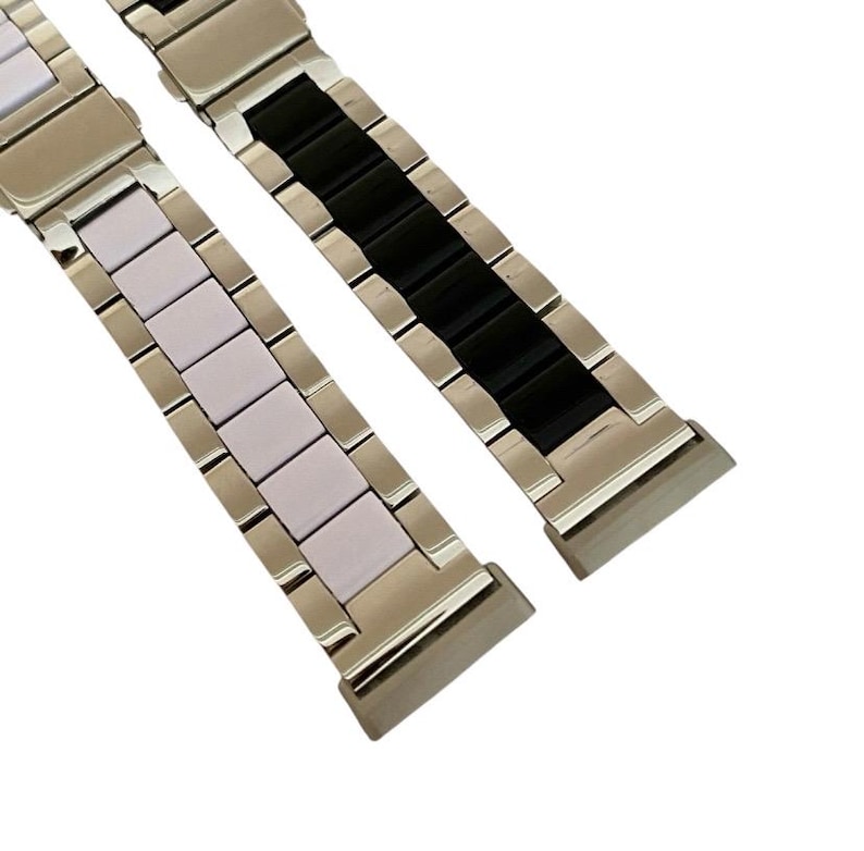 Puede incluir: Dos correas de reloj de metal con inserciones de diferentes colores. Una correa tiene inserciones blancas y la otra tiene inserciones negras.