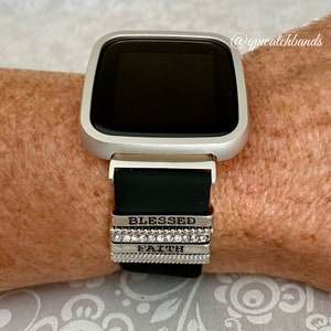 K&ouml;nnte beinhalten: Eine silberne Smartwatch mit einem schwarzen Silikonarmband. Das Armband hat drei silberne Charms mit den Worten "Blessed", "Faith" und eine Reihe von Strasssteinen.