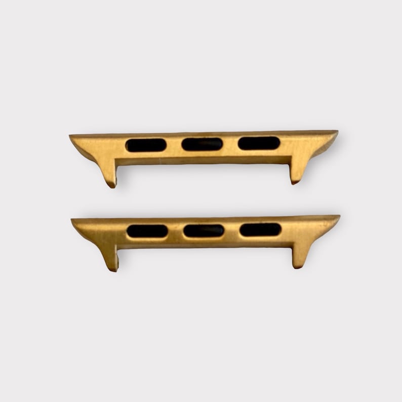 May include: Two gold-colored watch band adapters. Each adapter has three black rectangular holes and a curved, angled design on each end. The adapters are designed to connect a watch band to a watch face.