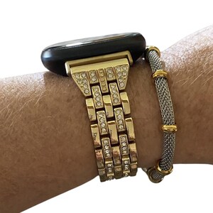 May include: Gold-toned watch band with embedded rhinestones and a black watch face. A silver mesh bracelet with gold accents is next to the watch. The watch band is connected to a wrist.