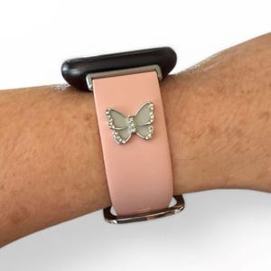 May include: A light pink watch band with a silver butterfly charm. The butterfly is adorned with small, clear crystals. The watch face is black, and the band is secured with a silver buckle.