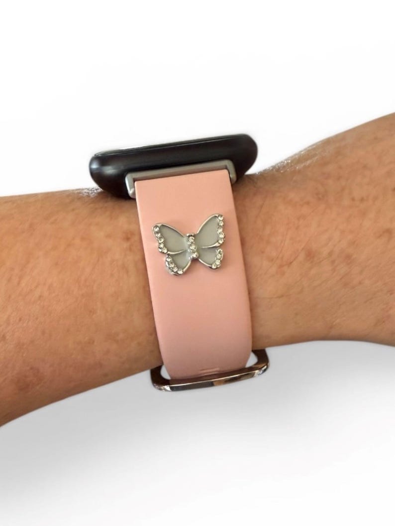 May include: A light pink watch band with a silver butterfly charm. The butterfly is adorned with small, clear crystals. The watch face is black, and the band is secured with a silver buckle.