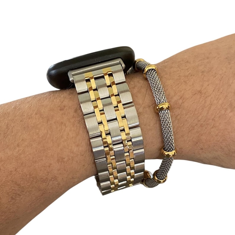 Peut inclure: Un bracelet de montre en m&eacute;tal argent&eacute; et dor&eacute; est attach&eacute; &agrave; une montre intelligente noire. Un bracelet en maille argent&eacute;e avec des accents dor&eacute;s est port&eacute; au poignet. Le bracelet de montre a un design &agrave; maillons rectangulaires avec des maillons centraux dor&eacute;s.