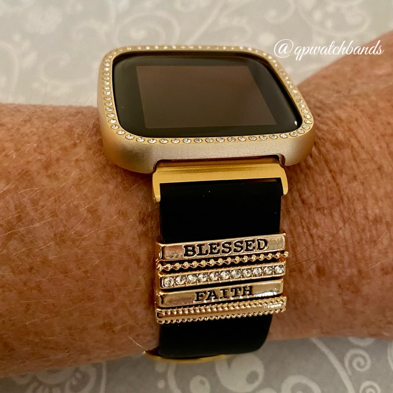 K&ouml;nnte beinhalten: Goldene Armbanduhr mit schwarzem Silikonarmband und goldenen Charms mit den Aufschriften "Blessed" und "Faith".