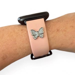 May include: A light pink watch band with a silver butterfly charm. The butterfly has a light blue center and is encrusted with small, clear crystals. The watch band is attached to a black watch face.