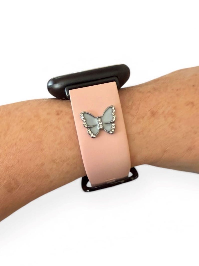 May include: A light pink watch band with a silver butterfly charm. The butterfly has a light blue center and is encrusted with small, clear crystals. The watch band is attached to a black watch face.