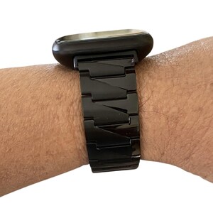 K&ouml;nnte beinhalten: Eine schwarze Smartwatch mit rechteckigem Zifferblatt und passendem schwarzem Metallarmband. Das Armband besteht aus verbundenen rechteckigen Gliedern. Die Uhr wird am Handgelenk getragen.
