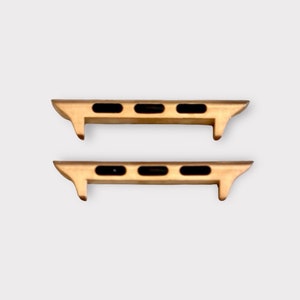 May include: Two gold-colored watch band adapters with black oval cutouts. The adapters have a rectangular shape with a curved edge and are designed to connect a watch band to a watch face. The background is a solid light gray.