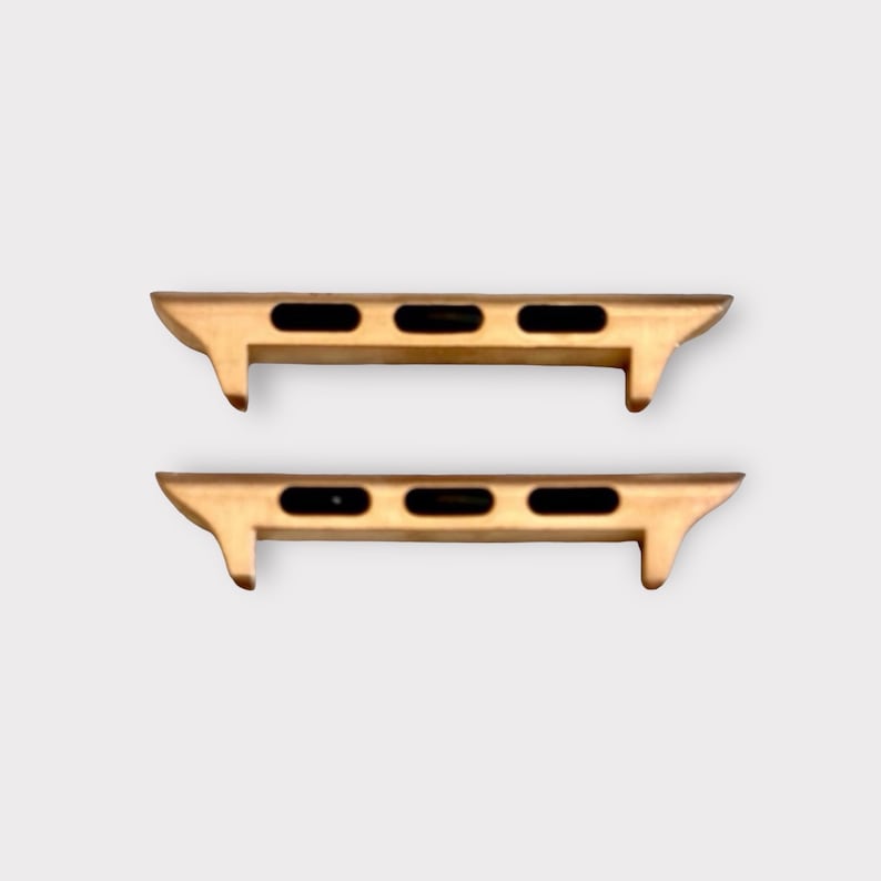 May include: Two gold-colored watch band adapters with black oval cutouts. The adapters have a rectangular shape with a curved edge and are designed to connect a watch band to a watch face. The background is a solid light gray.