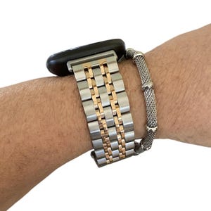Peut inclure: Un bracelet de montre bicolore avec des maillons argent&eacute;s et dor&eacute;s est fix&eacute; &agrave; une montre intelligente noire. Un bracelet en maille argent&eacute;e s&eacute;par&eacute; est &eacute;galement attach&eacute;. Les bracelets sont sur le poignet d'une personne.