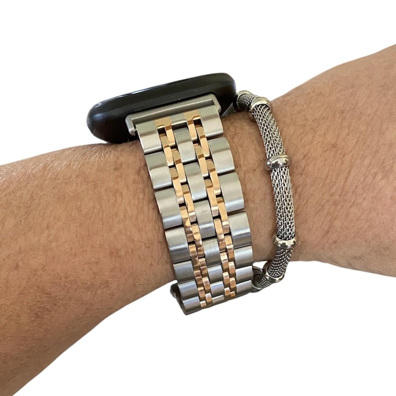 Peut inclure: Un bracelet de montre bicolore avec des maillons argent&eacute;s et dor&eacute;s est fix&eacute; &agrave; une montre intelligente noire. Un bracelet en maille argent&eacute;e s&eacute;par&eacute; est &eacute;galement attach&eacute;. Les bracelets sont sur le poignet d'une personne.