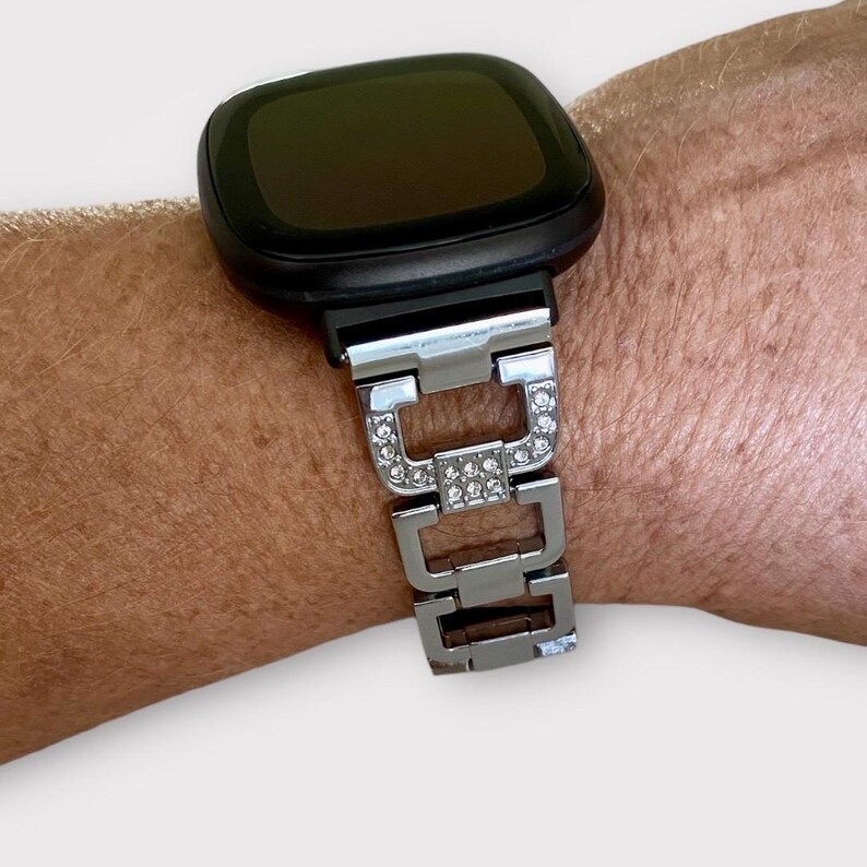 May include: A silver watch band with rhinestones, designed for a smartwatch. The band is made of metal links with a polished finish.