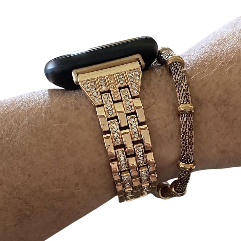May include: Rose gold watch band with a rectangular link design, embellished with clear rhinestones. A matching rose gold mesh bracelet is next to the watch band. The watch face is black.