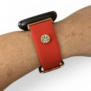 Peut inclure: Un bracelet en silicone rouge avec un accent rectangulaire noir et un fermoir dor&eacute;. Le bracelet est orn&eacute; d'un bijou rond en strass transparent. Le bracelet est port&eacute; sur un poignet.